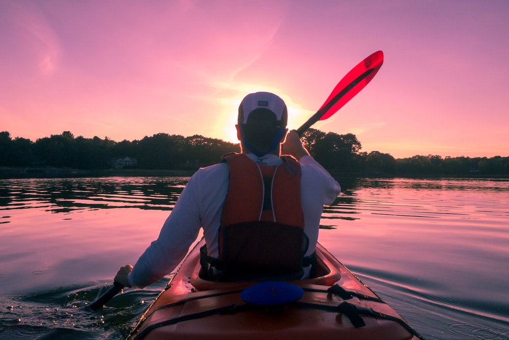 A man paddles a canoe on a calm lake at sunset. 