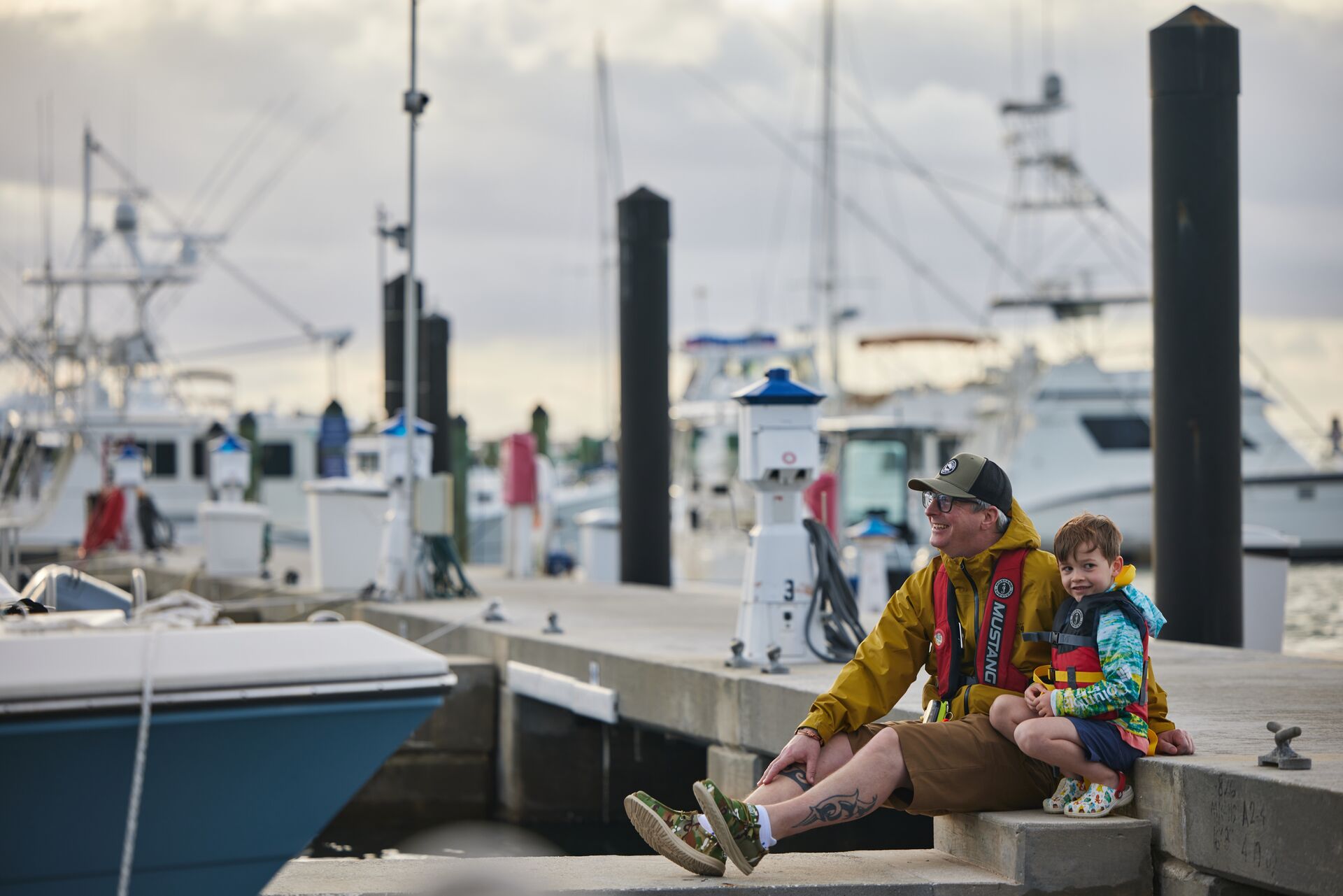 A smiling man and a boy sit on the dock of a marina while wearing life jackets. 