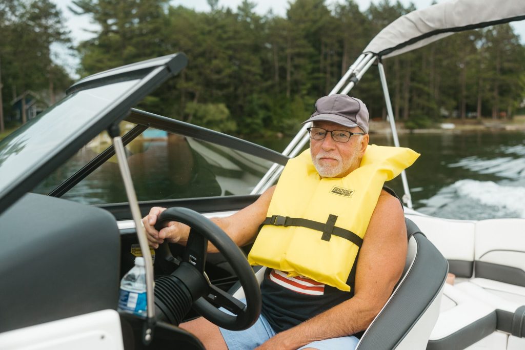 A man drives a boat on a lake while wearing a life jacket.