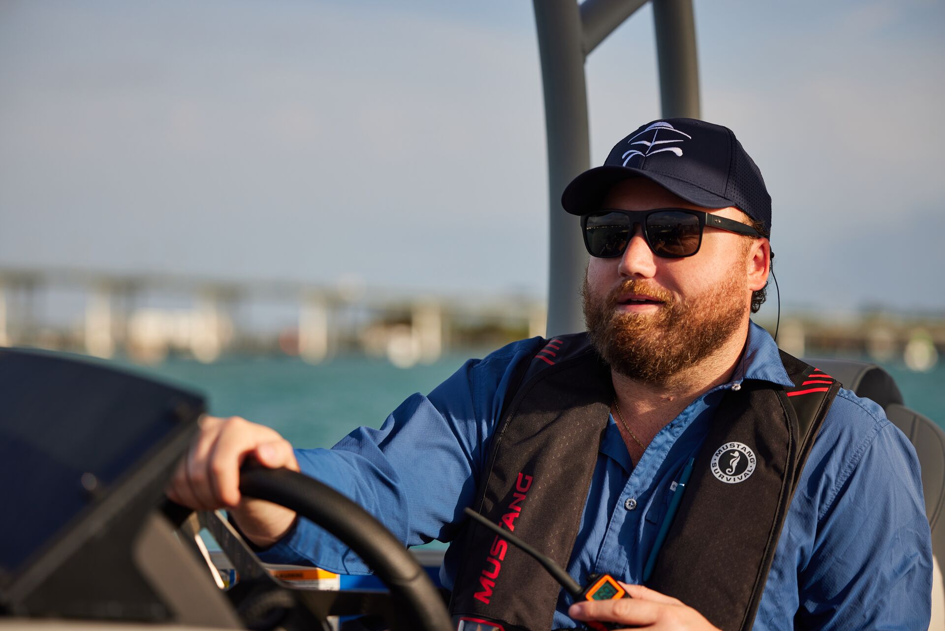 A man driving a boat while wearing a life jacket and holding a radio. 