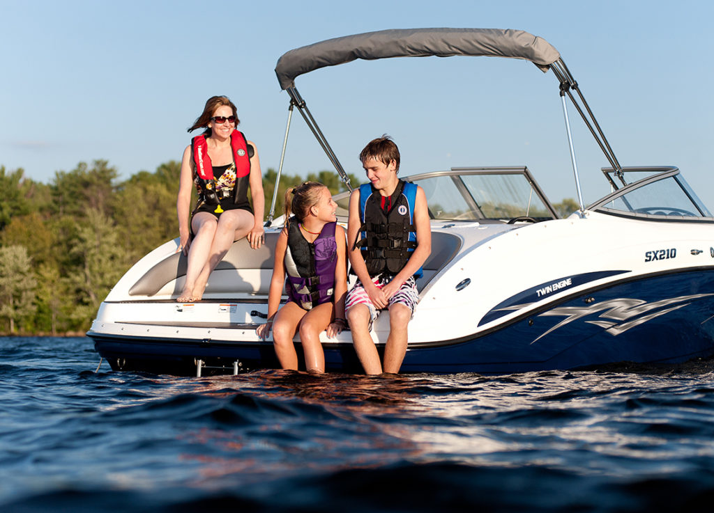 A woman and two kids wear life jackets while sitting on the back of a boat on the water.