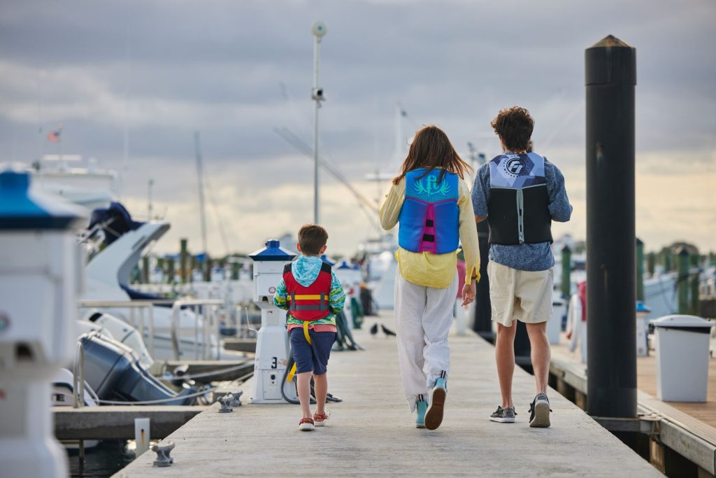 Kids wearing life jackets and walking near boats at a dock.