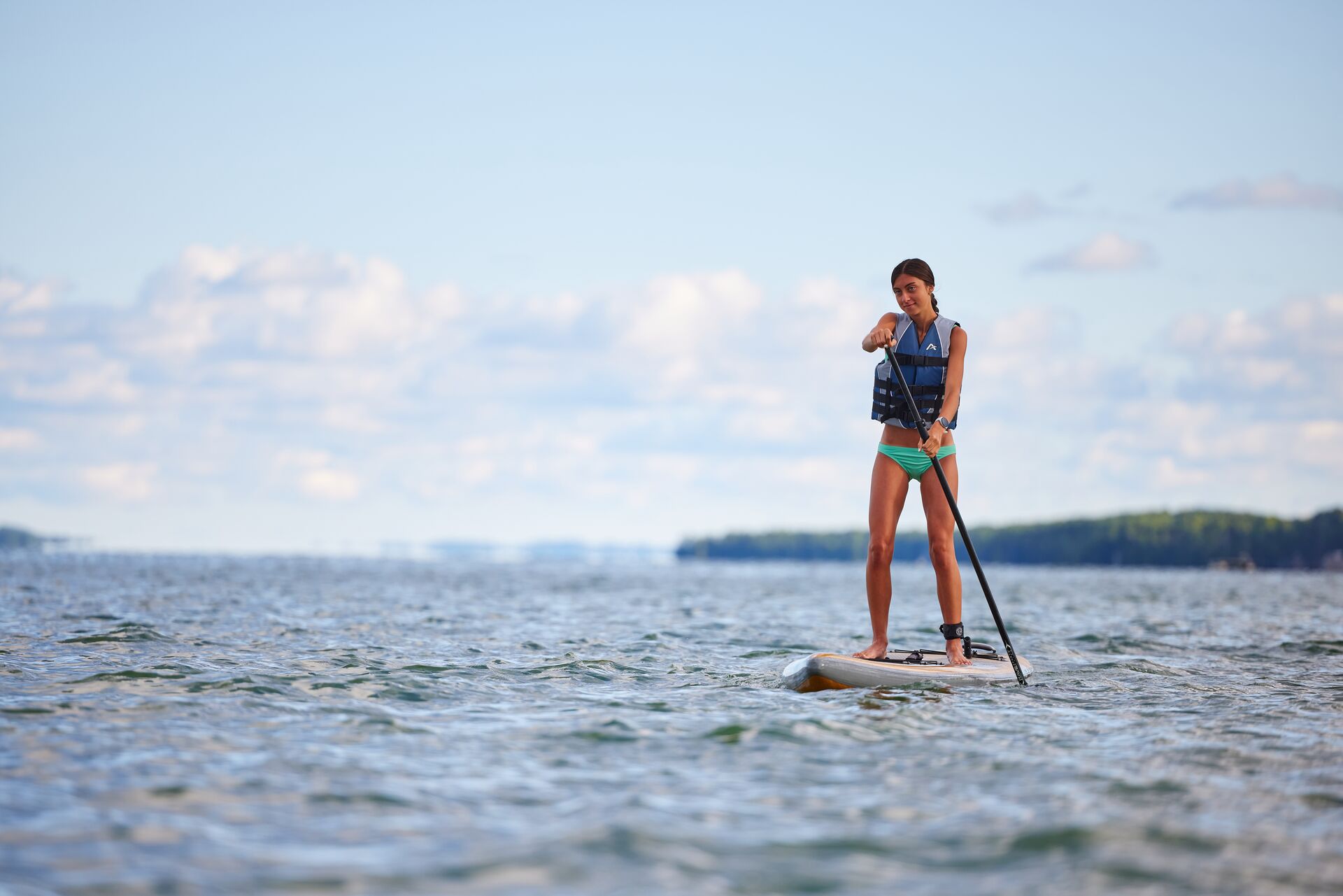 A girl stands on a paddle board with a paddle while wearing a life jacket.