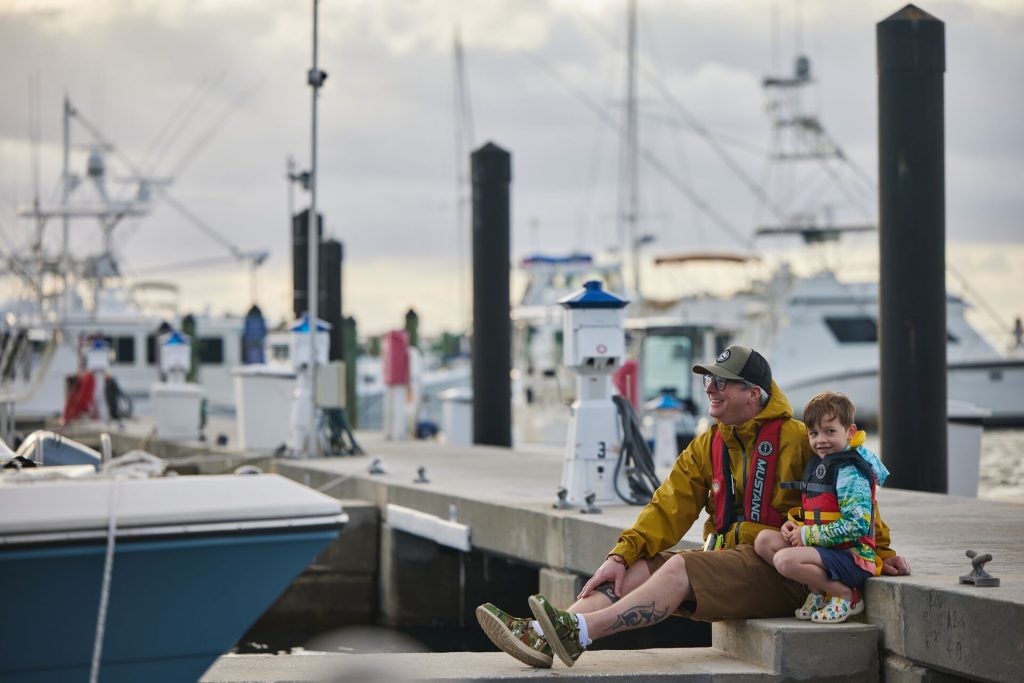 A man and boy in lifejackets smiling and sitting on the dock at a marina.