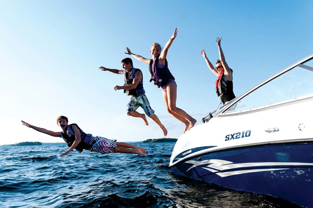 Kids jumping from a boat into the water while smiling and wearing life jackets.