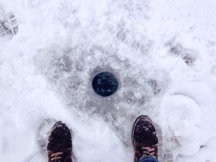 Two boot-covered feet standing next to a hole in the ice, ice fishing concept.