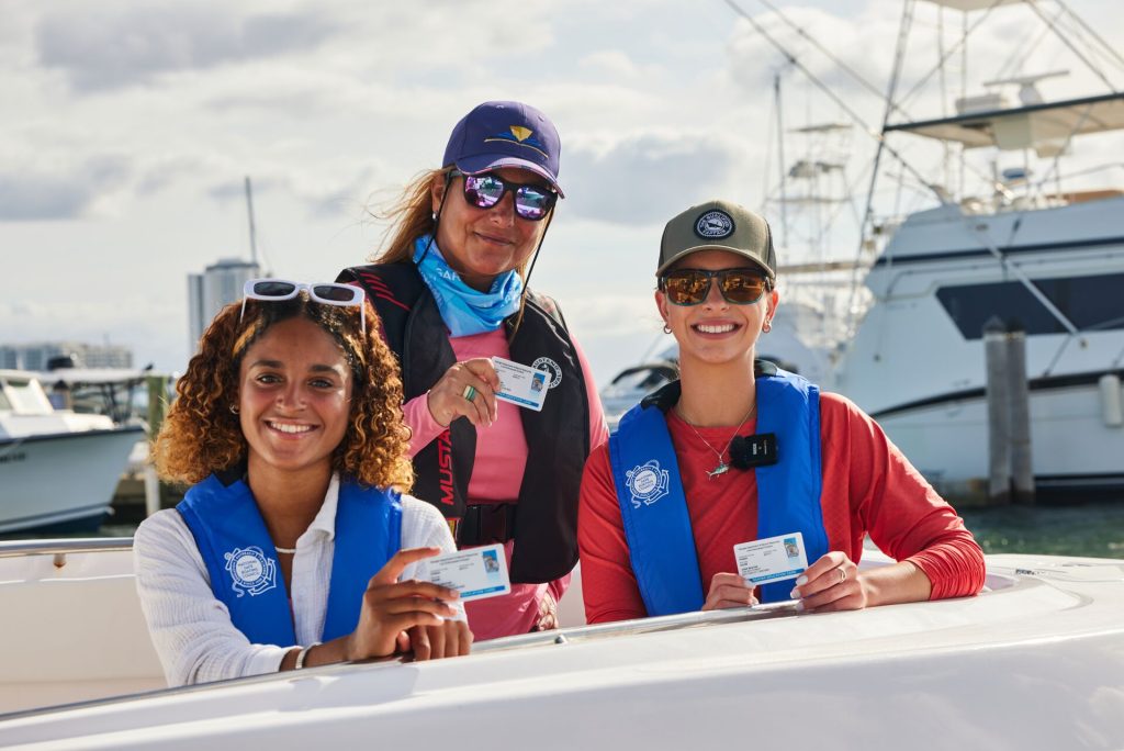 Three women holding boater education cards while smiling and wearing lifejackets on a boat.
